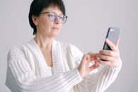 一位戴着眼镜、穿着白色毛衣的老年妇女正在室内使用智能手机