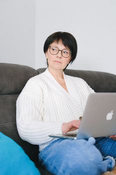 一位穿着羊毛衫的老妇人戴着眼镜，坐在沙发上使用笔记本电脑