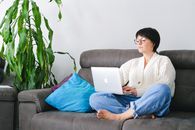 一位穿着羊毛衫的老妇人坐在沙发上，周围摆满了家居装饰，她正用着笔记本电脑