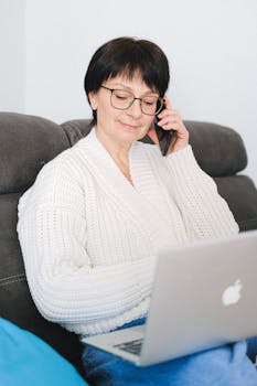 Elderly woman in a cozy sweater using a laptop and phone at home.