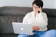 一位年长的妇女坐在沙发上，同时用着笔记本电脑和手机