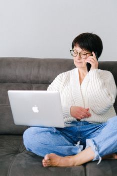 一位老年妇女坐在沙发上，同时用笔记本电脑和手机处理多项任务