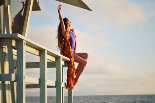 Full body side view of young slim beautiful female in purple swimwear and orange pareo chilling on tall wooden pier next to blue endless ocean under evening sky