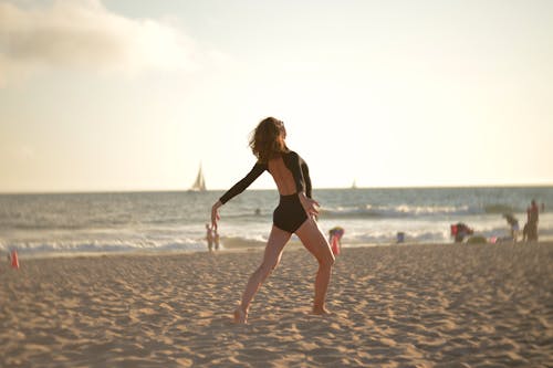 Full body back view of young expressive slender female dancer performing on sandy coast of wavy blue endless ocean in sunlight