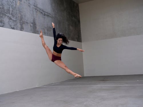 Full body of young concentrated ballerina in black crop top and purple underwear raising arms and outstretching legs while performing in simple studio with grey walls