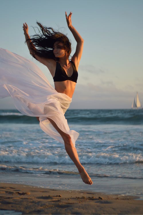 Full body of young focused ballerina in black bra and long white flying skirt raising arms and looking away while dancing on sandy shore of ocean under light blue sky