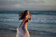 Side view of young happy female tourist with long dark hair in swimwear and pareo smiling and running on beach towards waving ocean during summer holidays