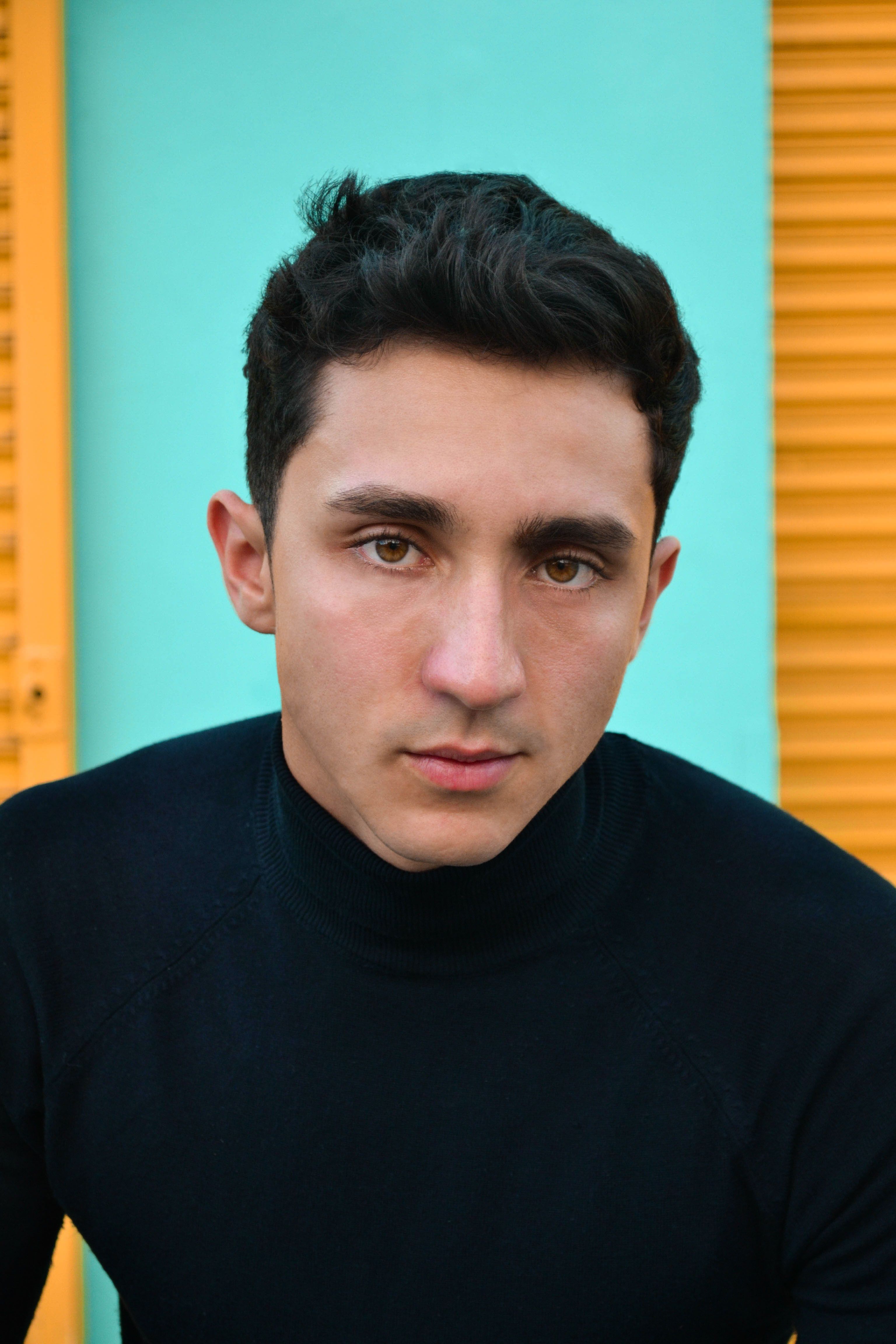 Close-up portrait of a man wearing a black sweater against a vibrant teal and yellow background.