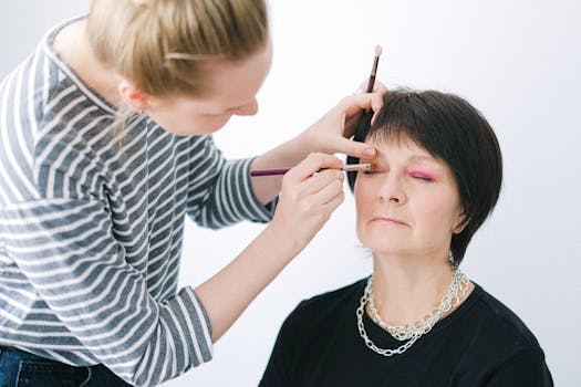A makeup artist applies vibrant eye makeup on a senior woman indoors.