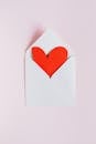 粉红色背景上，一个打开的白色信封顶视图，上面放着一颗手工制作的红纸心，作为情人节礼物