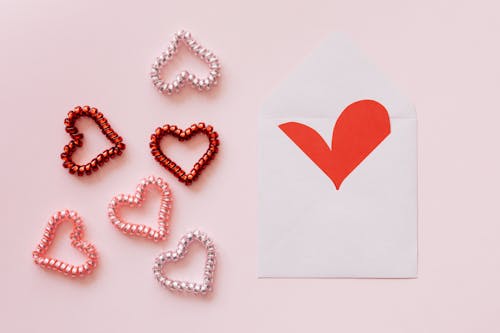 Top view of different hearts made of multicolored bobbles near white envelope with handmade red heart on pink background