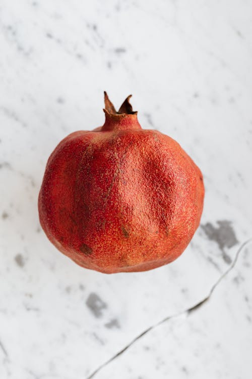 From above of whole ripe pomegranate with rough peel and small spiky leaves on top on marble surface with crack and grey spots