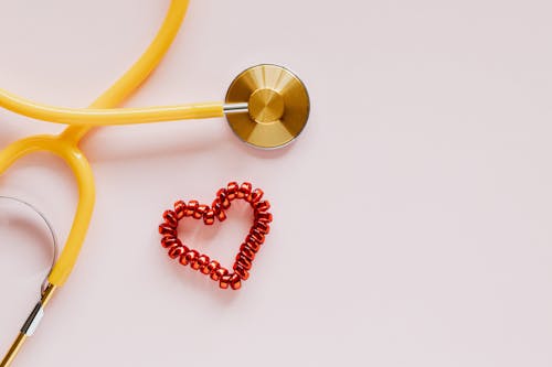 From above of yellow stethoscope near small red bead heart made for San Valentines Day on pale pink surface