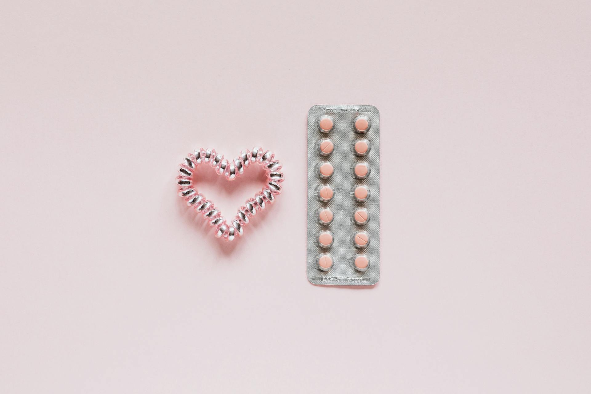 From above of whole blister of pills and shiny handmade heart of coil hair tie on pink surface