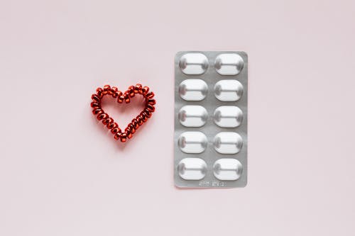 Top view of plastic blister with medical pills placed near red heart made of spiral elastic for hair on pink background