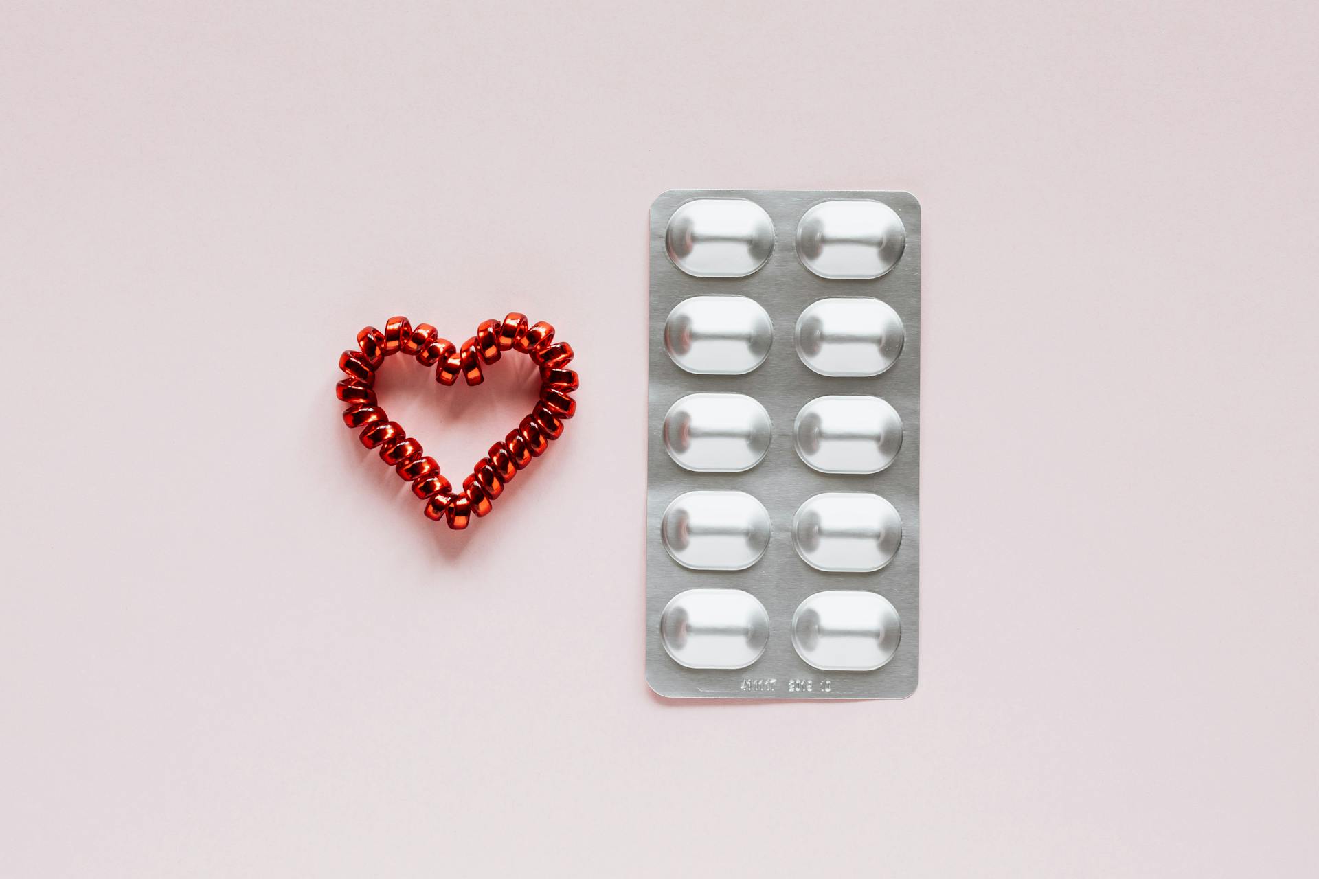 Top view of plastic blister with medical pills placed near red heart made of spiral elastic for hair on pink background