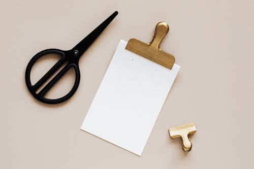 Top view of smal paper clip placed on beige surface near clipboard with gold clip and modern black scissors
