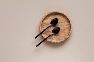 Top view of composition of round wooden plate with smooth surface and similar black metal spoons on beige surface