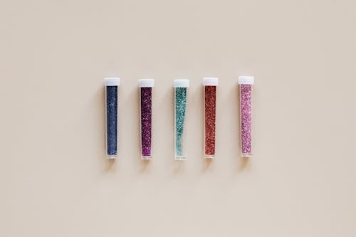 From above of transparent narrow plastic tubes with white lids filled with small multicolored shiny beads on beige background