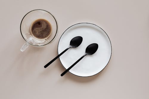 From above composition of ceramic plate with black spoons placed near glass cup of coffee on beige table