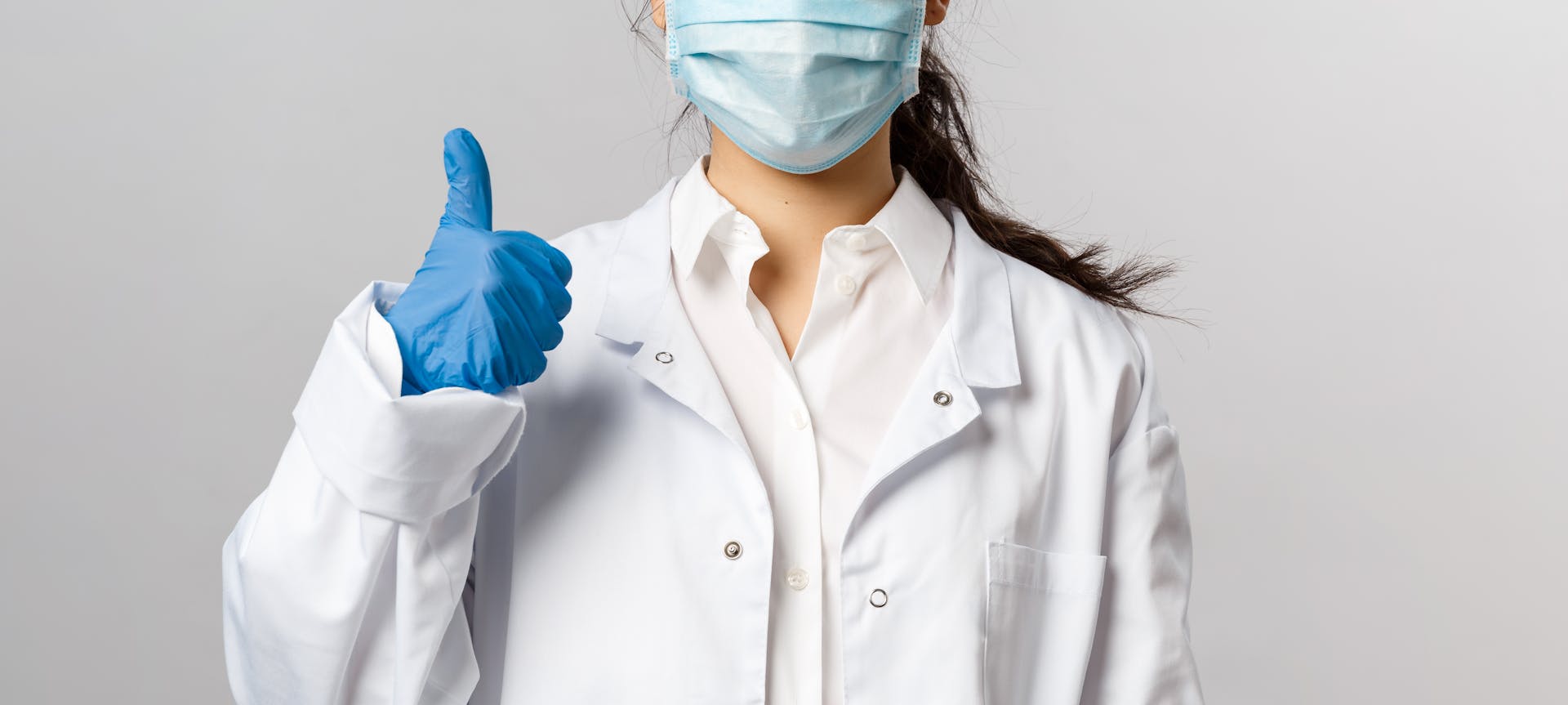 Healthcare worker in lab coat and mask giving thumbs up gesture.