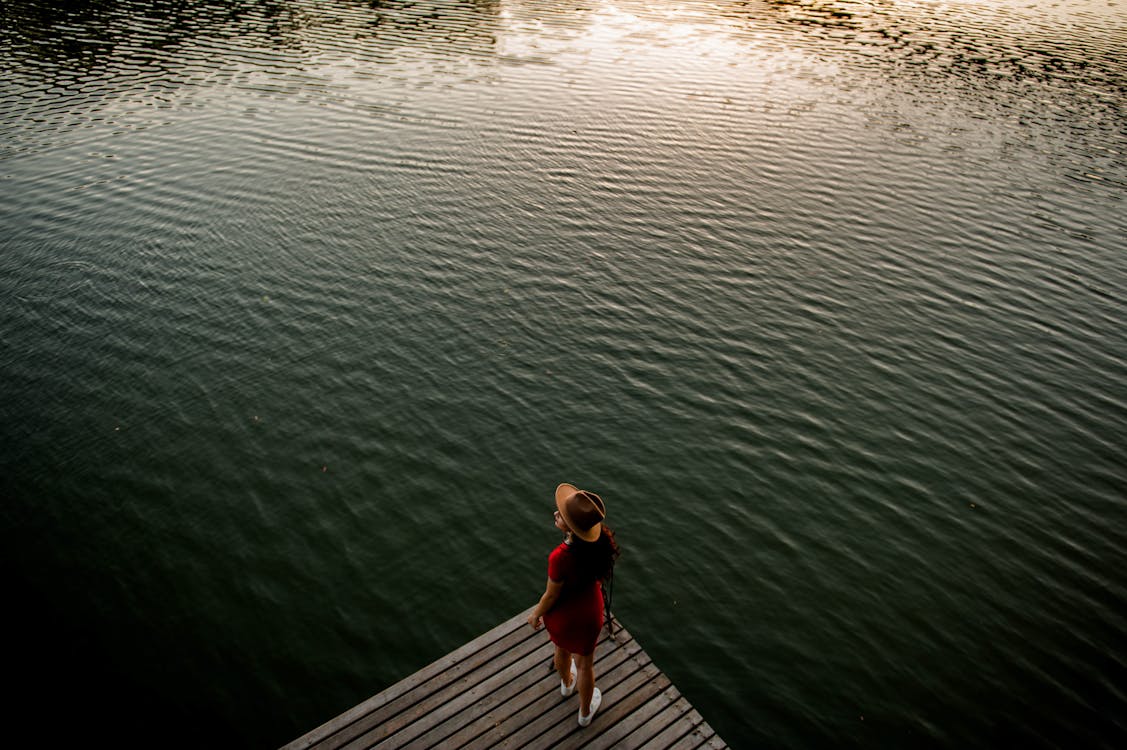 From above of anonymous female traveler in stylish dress and hat standing on wooden pier against rippling lake and admiring nature at sunset