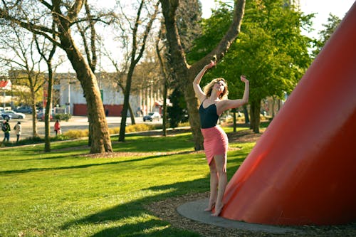 Young barefoot female dancer standing with raised arms near red construction and grass lawn in back lit while looking up
