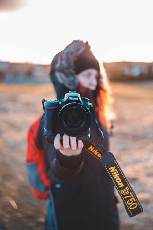 Side view of teen with backpack demonstrating modern style digital photo camera while looking away in back lit