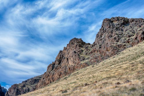 From below of picturesque sky with clouds over rough ridge and dry terrain with grass