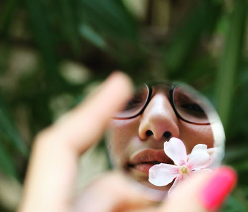 Crop female in eyeglasses looking in small round shaped mirror with flower in garden in summer