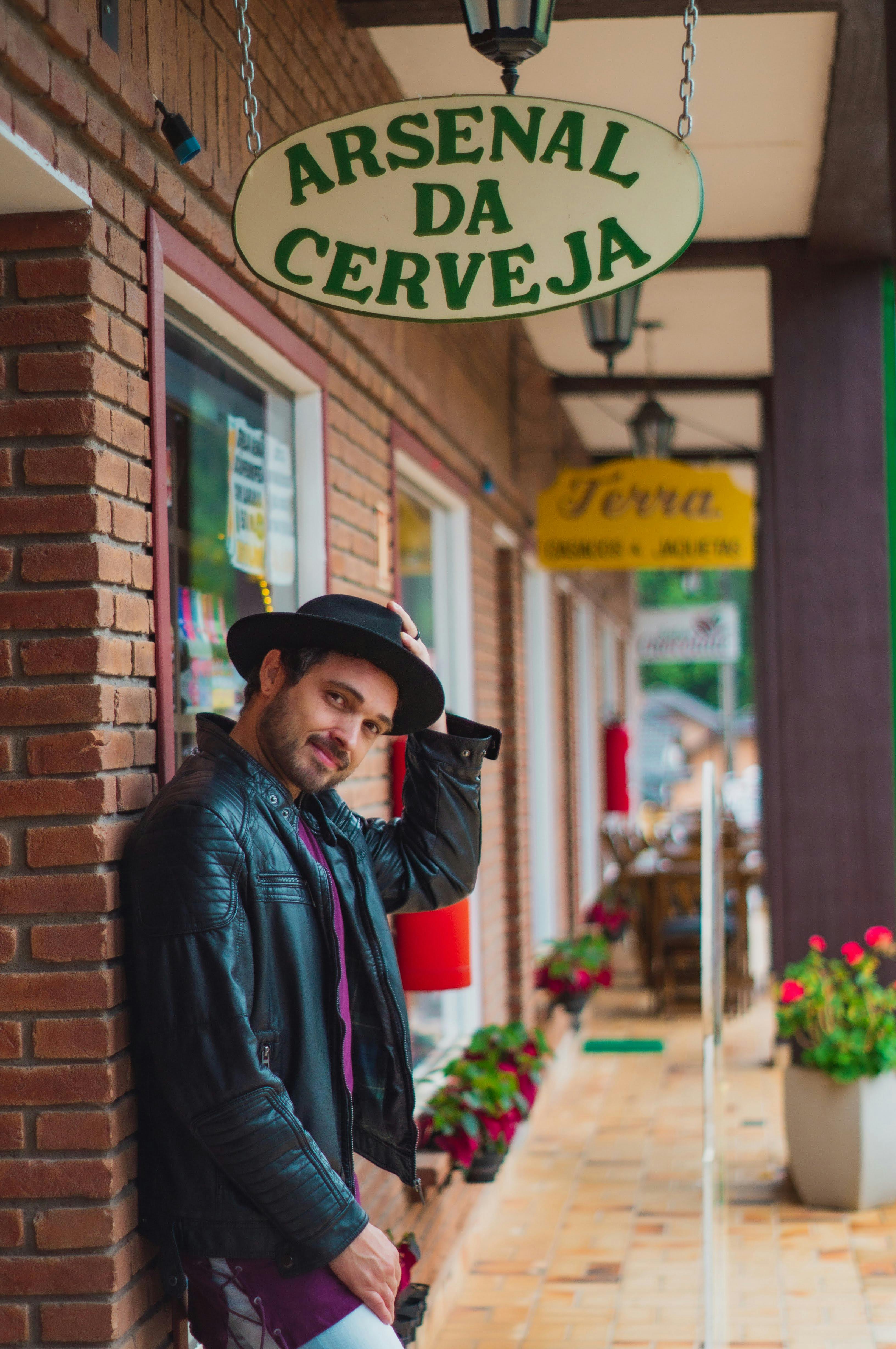 Side view of masculine bearded ethnic male in trendy apparel touching hat under signboard near house wall and looking at camera