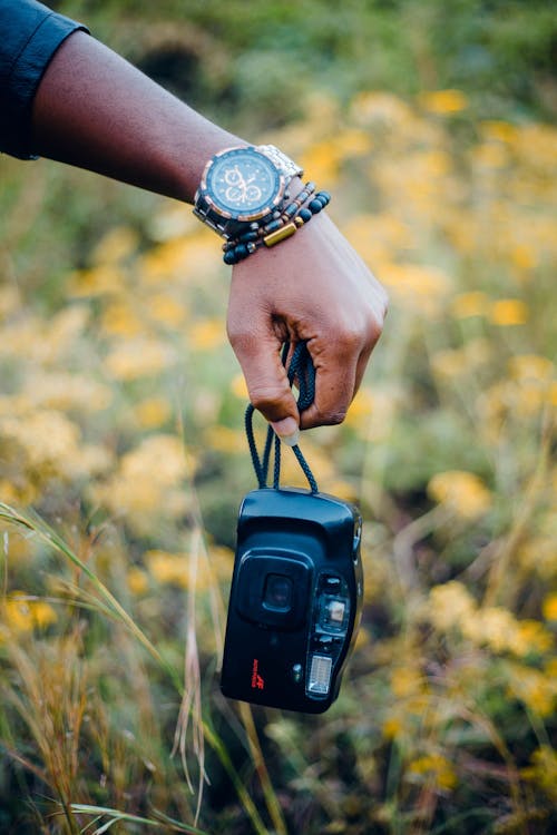 Crop anonymous female in accessories demonstrating professional photo camera in field with blooming flowers