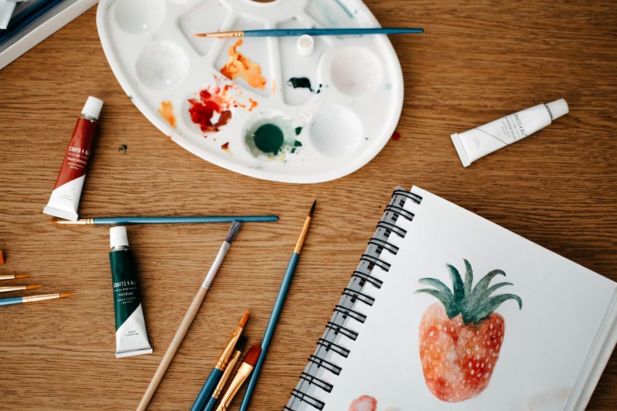 A watercolour palette, brushes and a sketchbook on a wooden desk
