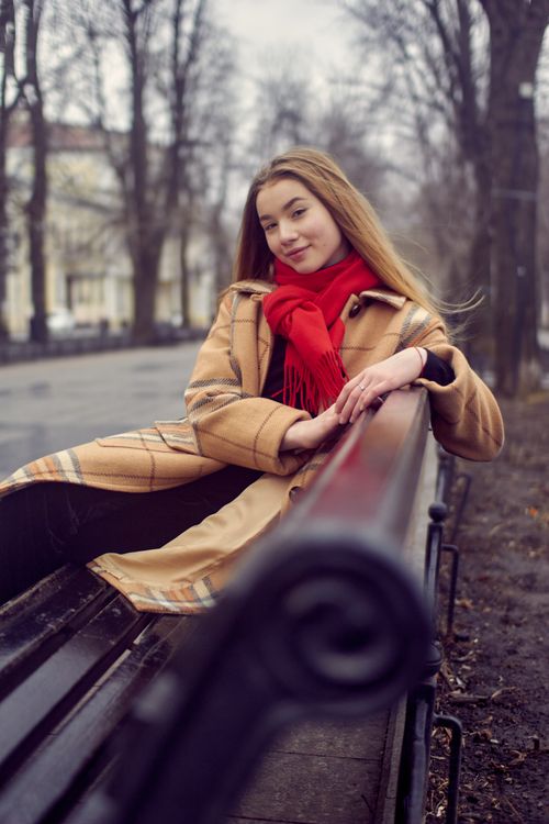 Free Caucasian woman wearing plaid coat and red scarf sitting on a bench in a winter park with bare trees. Stock Photo