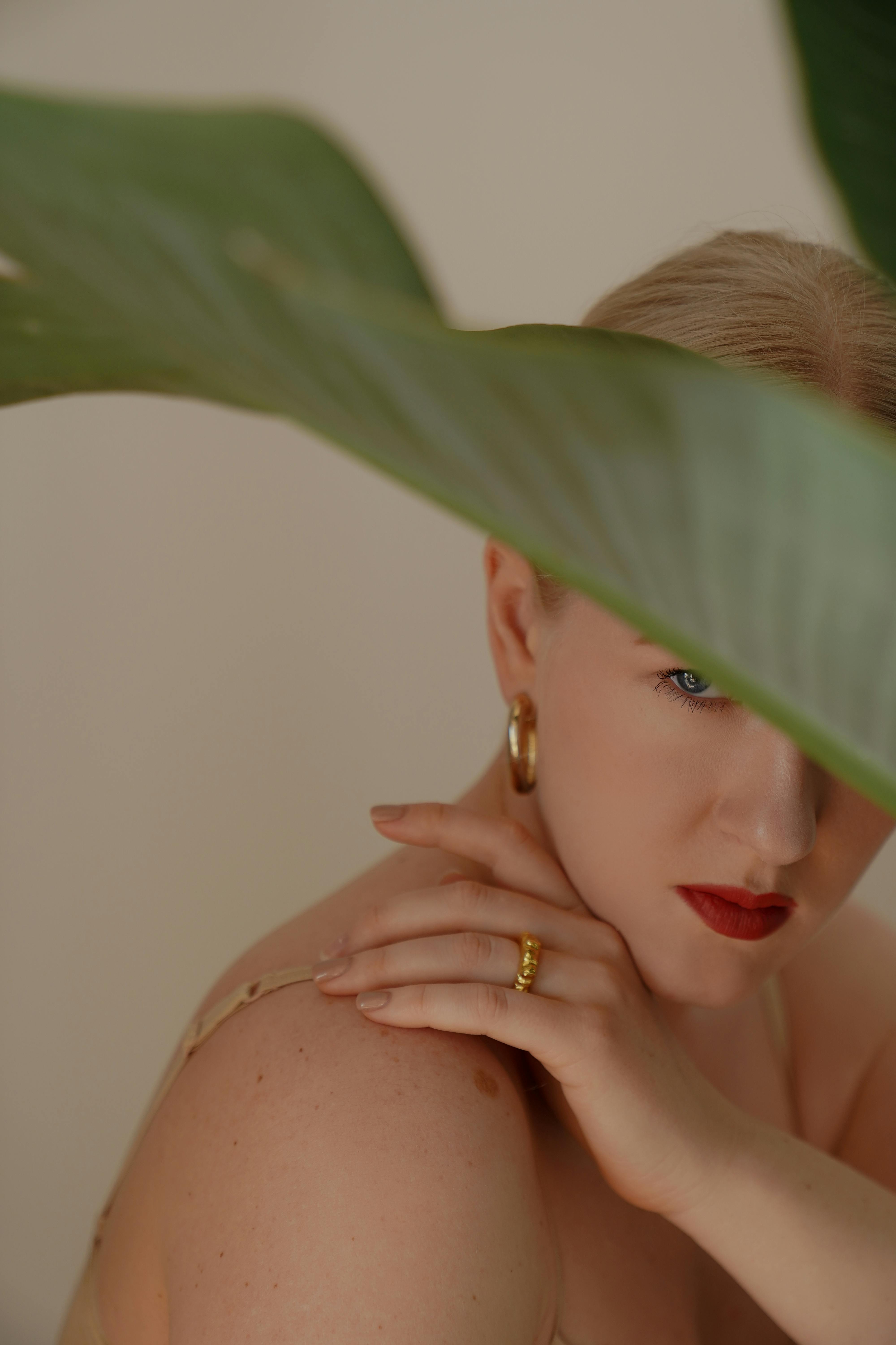 From above side view of adult serious blond lady in golden jewelry and beige underwear touching strap while looking away from under plant leaf