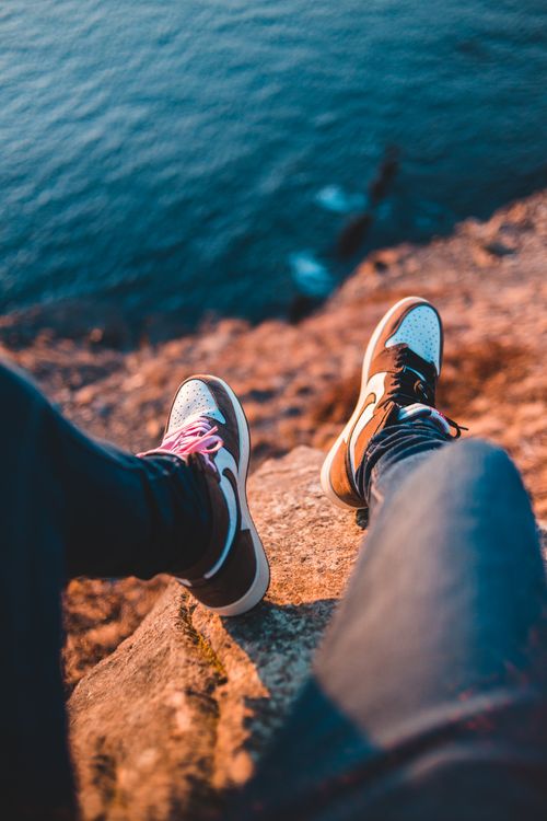 Free From above of crop anonymous traveler in jeans and sneakers sitting on bristly cliff with uneven surface above rippled sea in sunlight during vacation Stock Photo