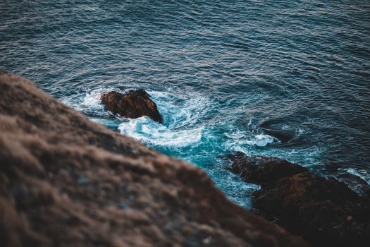 Spectacular from above view of rough brown mountain and foamy ocean with ripple surface creating swirl effect near stone in daylight