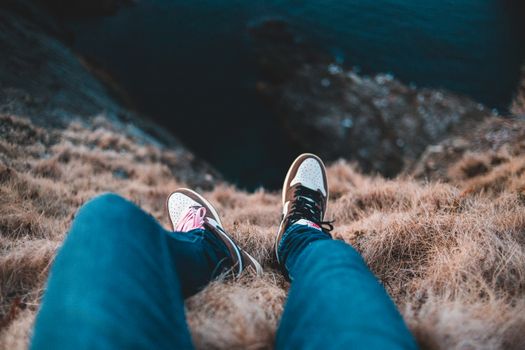 Top view of unrecognizable tourist in casual wear sitting on cliff with faded grass above rippled ocean in daylight during trip