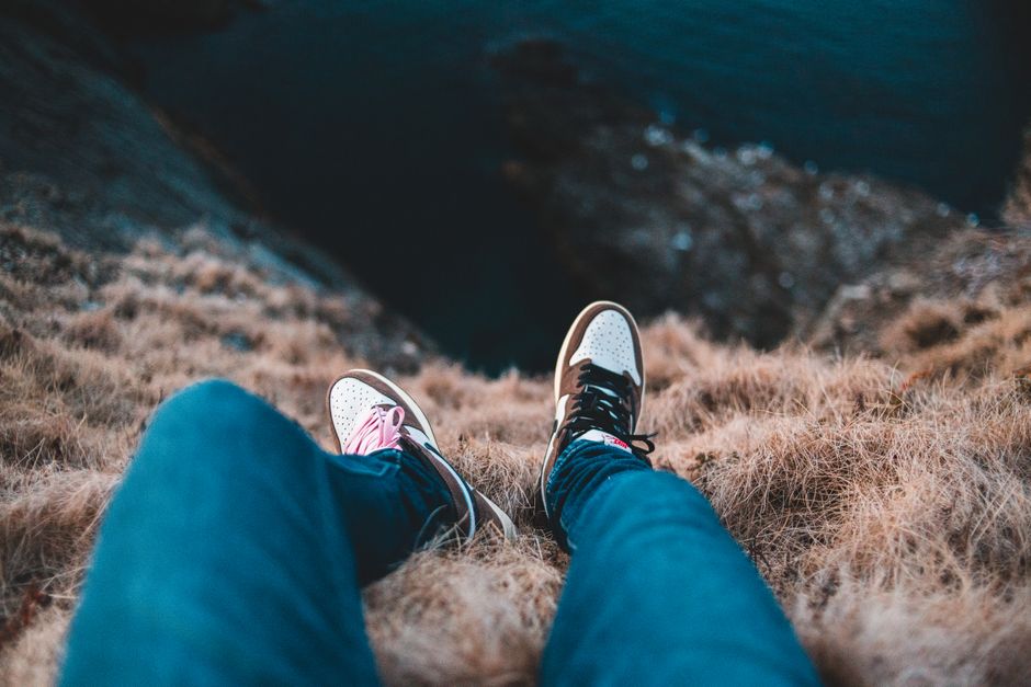 Top view of unrecognizable tourist in casual wear sitting on cliff with faded grass above rippled ocean in daylight during trip