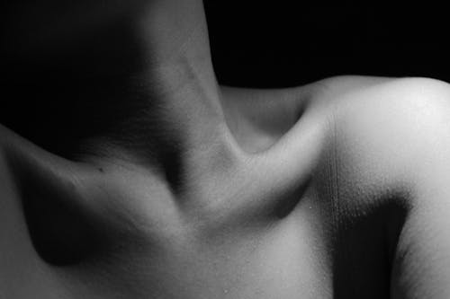 Black and white of crop unrecognizable slim nude person with pure skin on chest and shoulders with long neck and small mole above collarbones standing at home in darkness