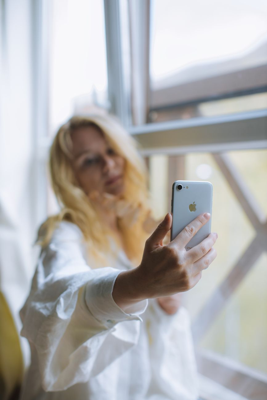 A woman with blonde hair takes a selfie by a window, embracing natural light indoors.