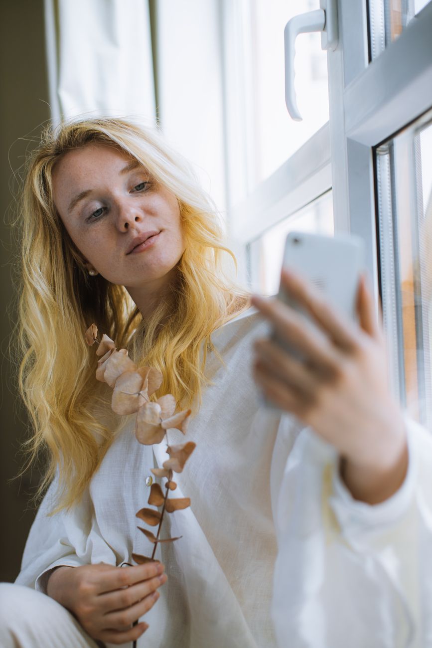 Blonde woman in white shirt taking a selfie indoors by window light.