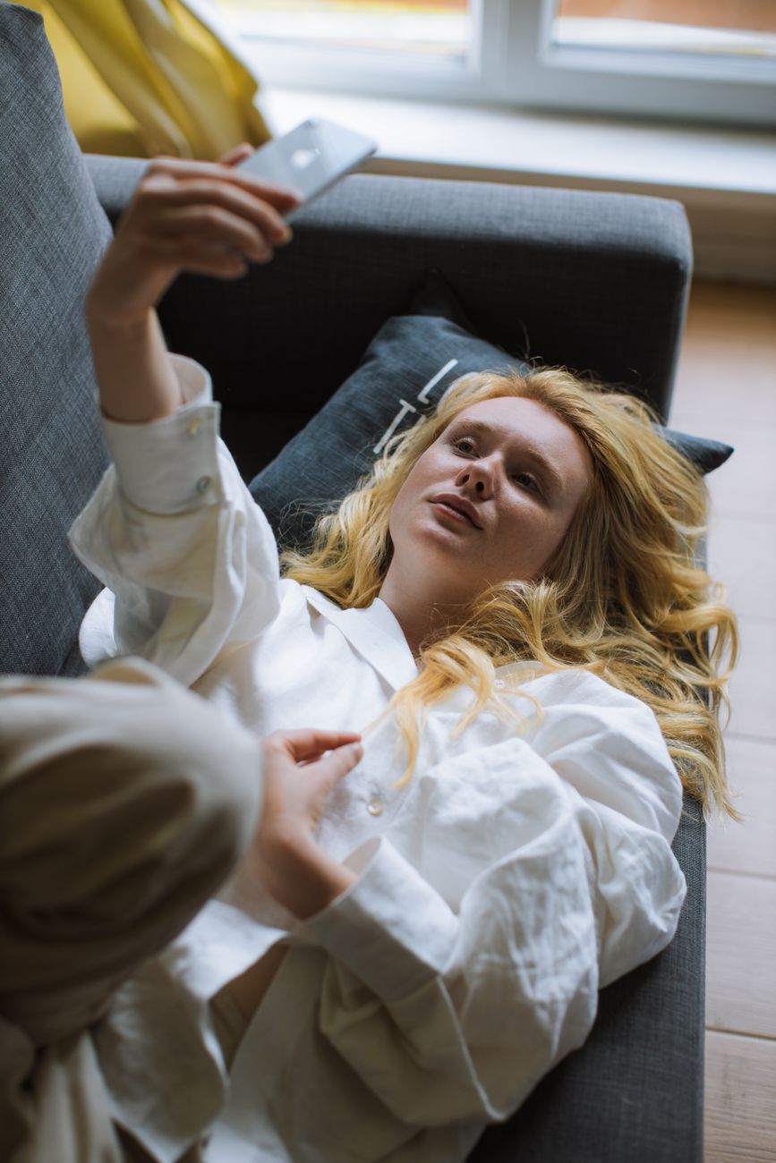 Blonde woman in white shirt taking selfie on sofa with smartphone.