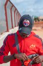 Serious African American male in casual wear and cap taking mobile phone out bag while standing in countryside near tall fence