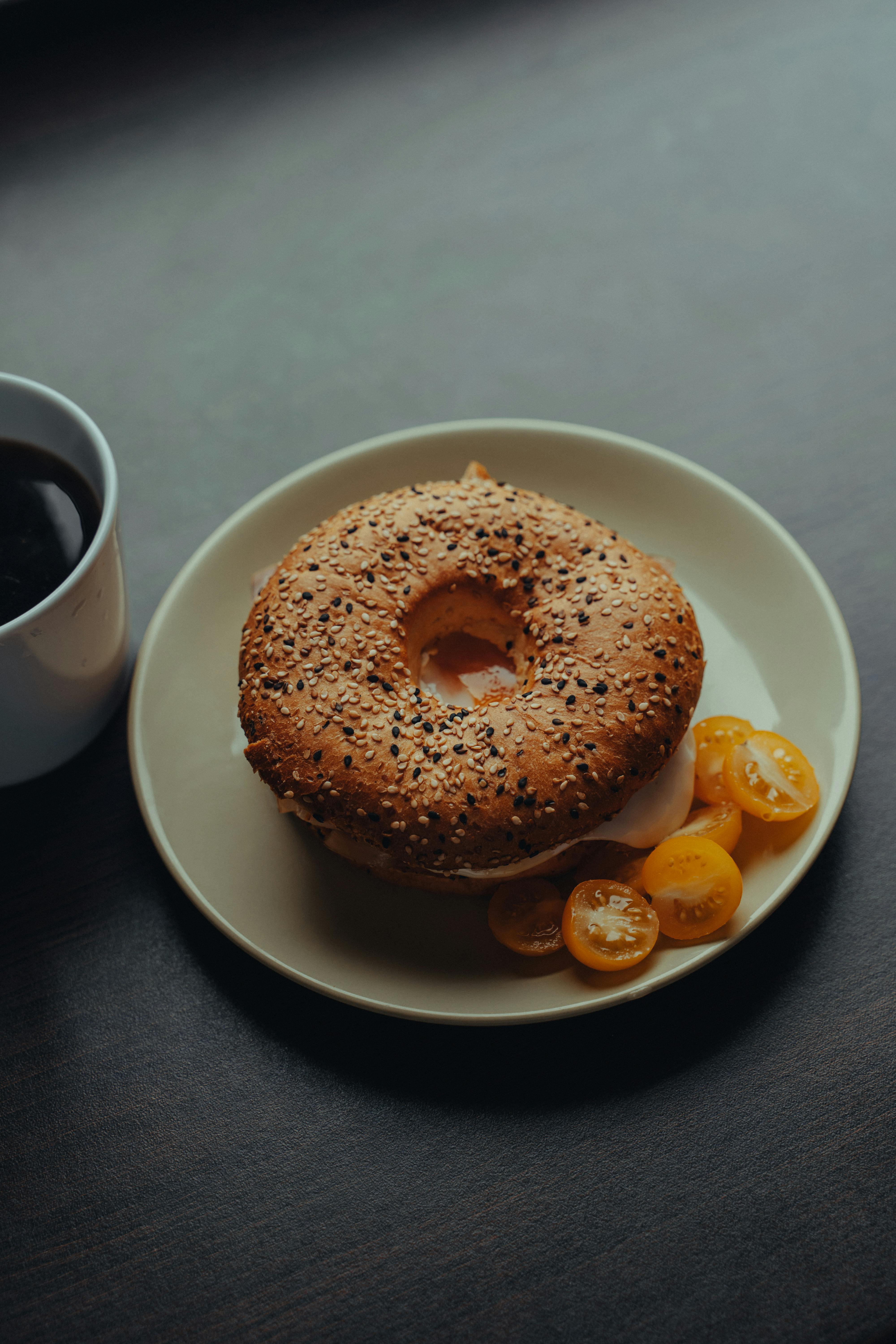 From above yummy bagel with fried egg served on white saucer with yellow cherry tomato and aromatic black coffee