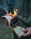 Crop anonymous male in casual wear holding burning book in flames while standing in evening forest