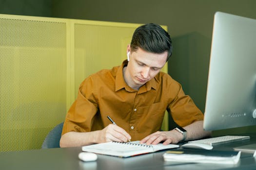 A man concentrating on his studies, taking notes at a desk with a computer.
