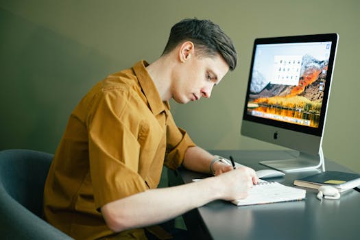 Adult male concentrating on notes with a desktop computer in a home office setting.