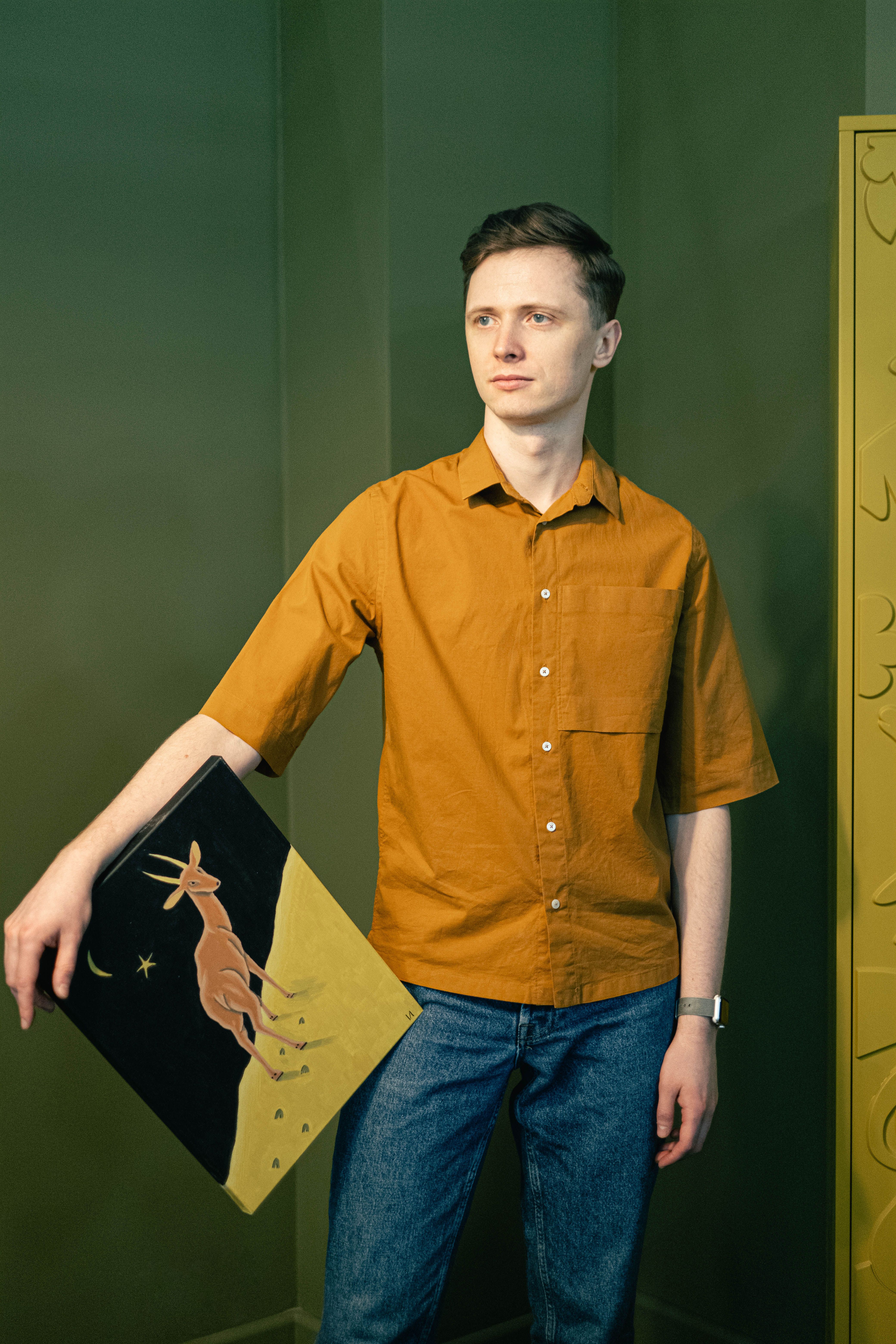 A young man in casual attire holding a modern painting indoors, focused and thoughtful.