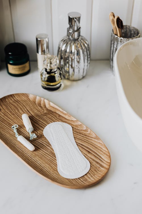 Free From above of cotton tampons and daily liner on wooden stand and toothbrushes and hand cleaner in jar and creams jars and perfume bottle placed on white marble surface near sink Stock Photo